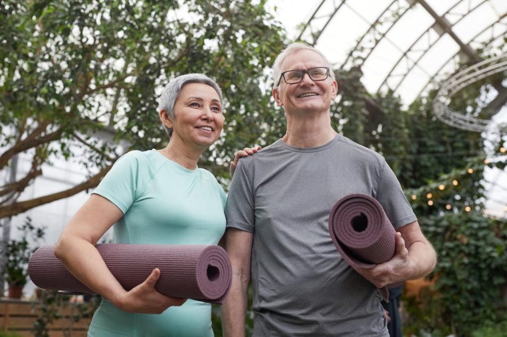 Happy senior couple holding yoga mats in a lush greenhouse setting.
